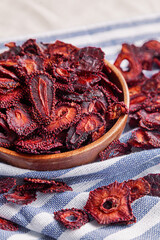 Rustic wooden bowl is filled with vibrant, deep red slices of dried strawberries, showcasing their intricate texture and natural beauty. The strawberries are arranged on a blue and white striped cloth