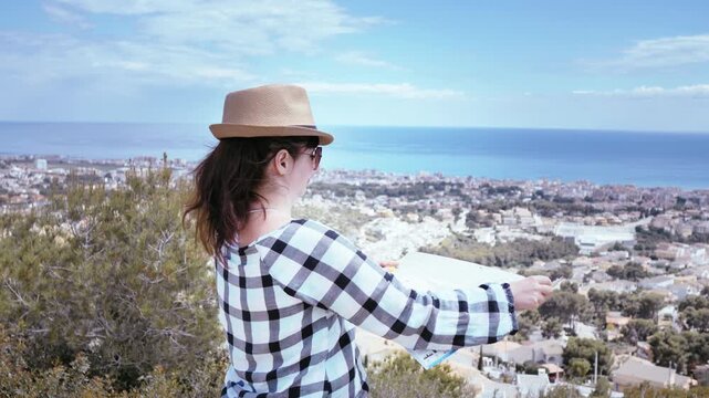 Adventurous Female Traveler Standing on a Scenic Hilltop, Gazing at a Coastal City Nestled by the Sea, While Thoughtfully Searching for Her Next Destination on a Detailed Map