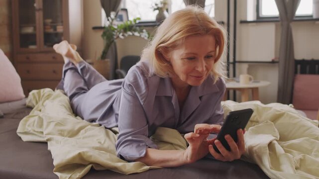 Portrait of attractive senior Caucasian woman with blond hair wearing silk lavender pyjamas relaxing on bed in morning chatting with friend in social net on smartphone