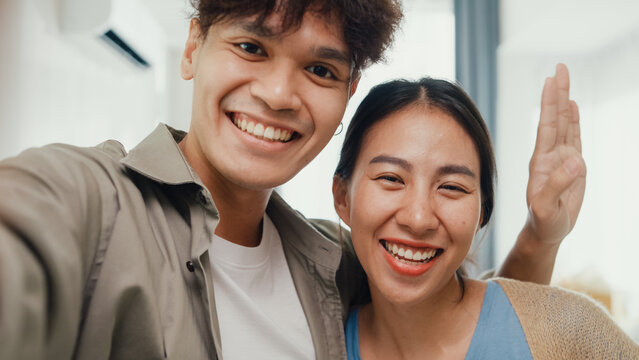Young happy Asian sweet couple video call look at camera smiling so proud and show off new house to their friends in living room. Husband wife move new home, Apartment Rent And Ownership Concept.