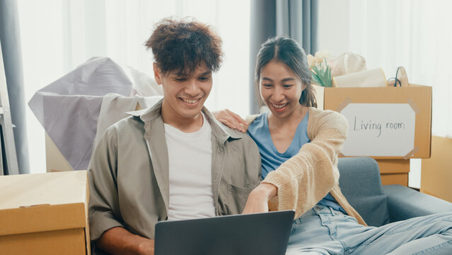 Young happy Asia couple sit on sofa couch use computer laptop online shopping furniture from decoration store in living room at home. Husband wife move new house, Apartment Rent And Ownership Concept.