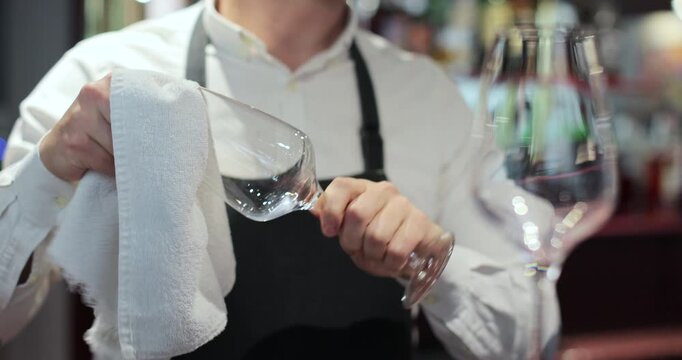 Close-up of male waiter polishing glasses at bar in restaurant. Hospitality industry, happy barista and wipe glassware for disinfection. Bar and cafe concept.