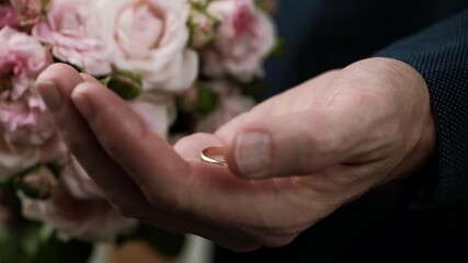 close-up nervous groom hand slowly opening to reveal golden wedding ring in his palm, with beautiful pink rose bridal bouquet out focus in background before ceremony.