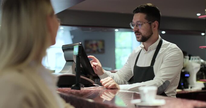 Young waiter with glasses in apron typing on touch screen cash register working and talking with woman at bar or restaurant.