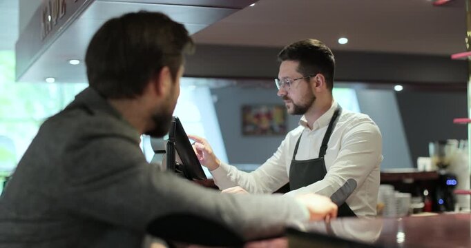Young waiter with glasses in apron typing on touch screen cash register working and talking with man at bar or restaurant.