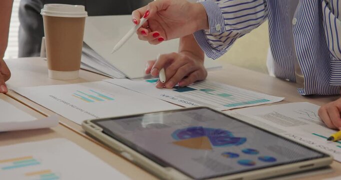 Close-up view of diverse young business team collaborating with digital tablet and printed charts. Hands pointing at colorful graphs and analytics during a strategy planning session at office.