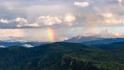Vibrant rainbow appears above forested mountains after summer rain