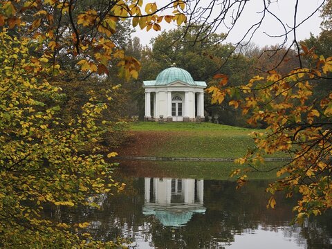 Temple on the Swan Island in the Karlsaue, surrounded by autumnal trees, whose reflection can be seen in the calm water, Kassel, Hesse, Germany