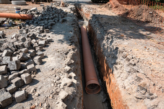 Instala&ccedil;&atilde;o de tubula&ccedil;&atilde;o corrugada numa valeta de terra para drenagem ou saneamento b&aacute;sico, com pilhas de pequenos paralelep&iacute;pedos ao lado a Indicar obras de infraestrutura urbana