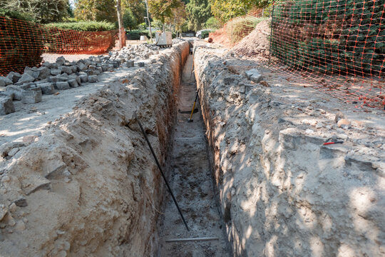 Vala profunda e estreita escavada numa &aacute;rea urbana para obras de infraestrutura de saneamento com o solo removido e empilhado nas laterais e algumas ferramentas espalhadas na trincheira