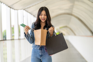 Asian young woman opening shopping bags excited with new purchases at outdoor mall, stylish casual outfit, joyful shopping experience, retail therapy, trendy fashion consumer lifestyle modern urban