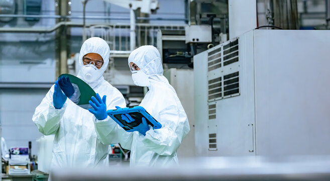 Male and female workers in protective clothing inspecting silicon wafers while looking at a tablet in a semiconductor factory