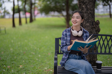 Woman student sitting on a park bench, holding an open book and pen, wearing headphones around her neck, smiling and looking away, surrounded by green trees and grass