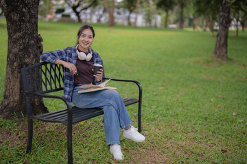 Young Asian woman sits on a park bench with headphones around her neck, smiling as she reads a book and sips coffee, enjoying a peaceful campus afternoon outdoors