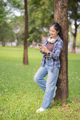 Young Asian woman posing with a casual outfit and headphones around her neck, smiling cheerfully while browsing on her smartphone, leaning on a tree trunk in a lush green park