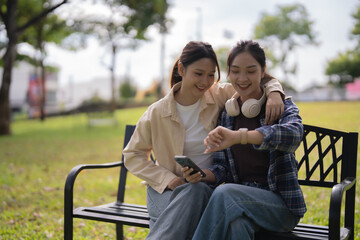 Two happy young women friends sitting on a park bench, looking at a smartwatch and holding a mobile phone, representing friendship, connectivity, and modern lifestyle outdoors