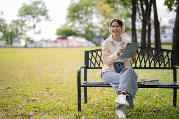 Fototapeta premium Young Asian woman wearing headphones and using a digital tablet with a stylus while sitting on a park bench, engaging in e-learning in a serene outdoor environment