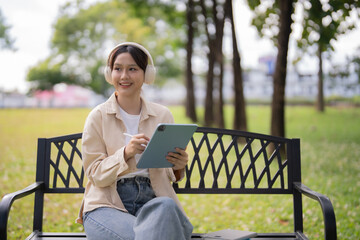 Fototapeta premium Young Asian woman in headphones smiling while working or studying outdoors on a park bench, using a tablet and stylus for remote learning, creative work, and relaxation