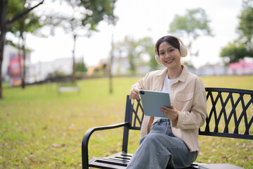 Fototapeta premium Young Asian woman sitting on a park bench, happily engaging with her digital tablet and wearing wireless headphones, enjoying a moment of online learning or relaxation outdoors