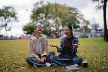 Fototapeta premium Two cheerful Asian female university students sitting on green grass, laughing and expressing joy while throwing white flowers during a relaxing time in a campus park