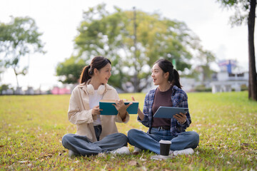 Two smiling young Asian female college students are sitting on green grass in a park, engaging in a lively discussion while studying together with an ebook and a digital tablet