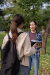 Fototapeta premium Two young Asian women smiling and talking outdoors on a university campus, one with a tablet and documents, the other with a backpack, sharing ideas and studying together in nature