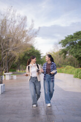 Asian female students walking together, happily engaging in conversation while holding a coffee cup and a book on a university campus path, embodying friendship and a casual lifestyle outdoors