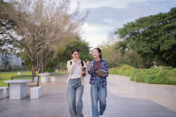 Young Asian female students walking and laughing along a university campus path, chatting between classes while holding books and coffee, relaxed and enjoying a sunny day