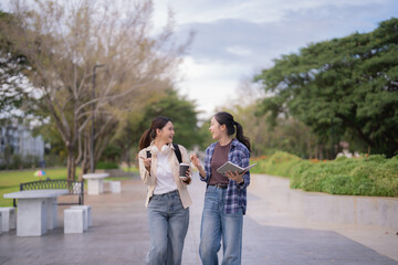 Fototapeta premium Two cheerful university students stroll across campus smiling and talking, carrying textbooks, backpacks and coffee as they enjoy a sunny break between classes and share a friendly moment