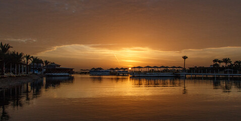 Serene sunset over calm water, reflecting vibrant orange and yellow hues, with silhouettes of palm trees and boats creating a tranquil coastal atmosphere. Egypt, Red sea, Hurghada