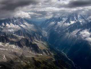 Aerial view of majestic snow capped mountain range rugged peaks deep valleys and rock formations under a dramatic cloudy sky with breaking sunlight