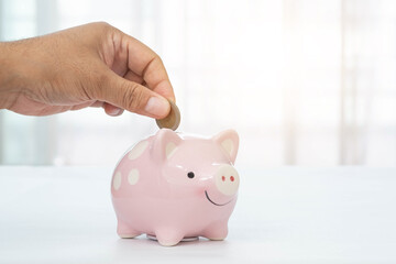 Man's hand putting money in piggy bank on white table surface and blurred background. Savings concept.