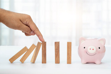 Man's hand pointing at falling wooden blocks to stop falling and piggy bank on white tabletop and blurred background. Financial and investment risk concept.