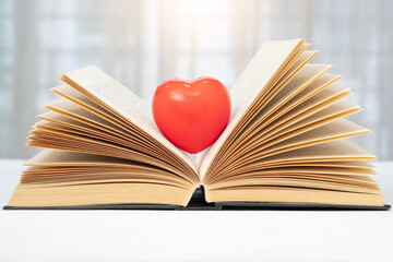 Red heart on open book side view on white table surface and blurred background.