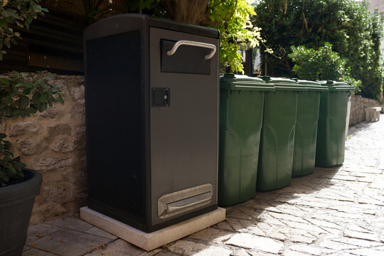 A tall, dark gray communal waste container stands on a stone path next to a line of standard green recycling bins, surrounded by lush foliage and stone walls.