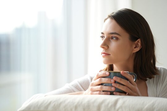Thoughtful young woman holding a warm cup of coffee by a bright window at home