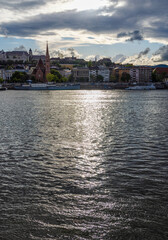 Obraz premium Scenic cityscape view across a calm river at sunset with sunlight reflecting off the water. Historic buildings and a church spire rise on the opposite bank under dramatic clouds