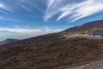 Expansive view of Mount Etna volcanic terrain under a bright blue sky. Rugged lava fields stretch across the slope, with a small settlement and parked cars visible in the distance.