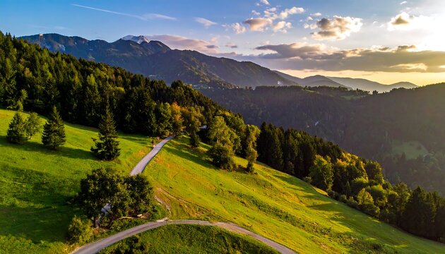 A sunlit mountain landscape. A winding road weaves through vibrant green hills and dense forests, leading towards distant peaks under a cloudy sky