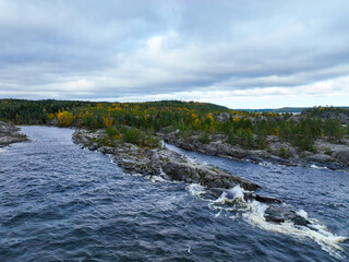 Aerial view of dark, turbulent water crashing over a narrow, rocky outcrop, separating two channels of water, with autumn forest on the distant shore