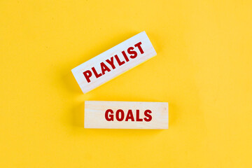 On a bright yellow background, two wooden blocks are placed showing the words PLAYLIST and GOALS, suggesting the act of creating musical experiences with intentional song choices