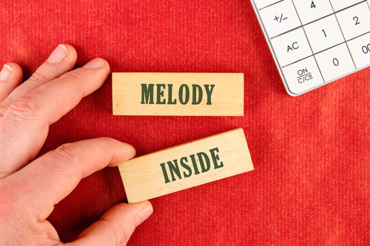 On a red surface, a hand holds a block with the word INSIDE, while another block reads MELODY. A calculator peeks into frame, hinting at a need to calculate the value of self