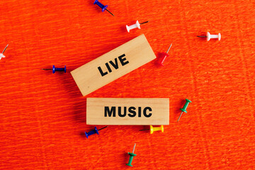 Two wooden blocks, one reads LIVE and the other MUSIC. They rest on textured orange paper, scattered with colorful pushpins. It suggests an announcement or invitation