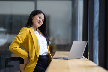 Young businesswoman experiencing back pain while working on a laptop in office, physical strain and health challenges associated with prolonged desk work.