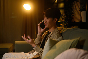 Casual Asian man enjoying a pleasant phone call at night, sitting comfortably in a softly lit living room