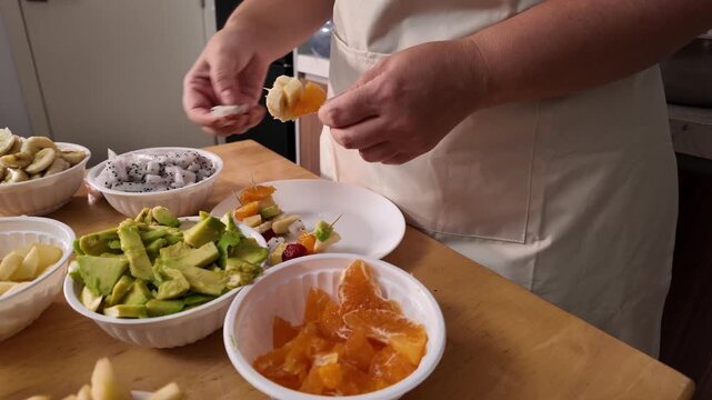 A woman's hands are making fruit kebabs, stringing pieces of fruit on a skewer