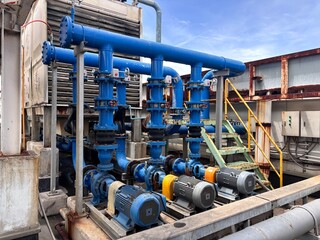 An intricate industrial cooling system on a rooftop featuring electric pumps, a network of blue pipes, flanges, and valves. This HVAC and plumbing machinery highlights heavy industry and utility maint