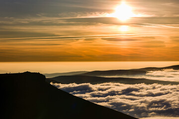 Silhouettes of hikers on Corn Du mountain peak watching a golden sunset over a sea of clouds