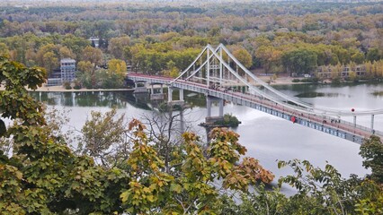 Suspension pedestrian bridge crossing a calm river surrounded by autumn trees. Concept of urban...