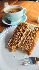Slice of layered carrot cake with cream and caramel served on a plate beside a cup of coffee. Concept of cozy cafe moment, dessert enjoyment, and sweet indulgence